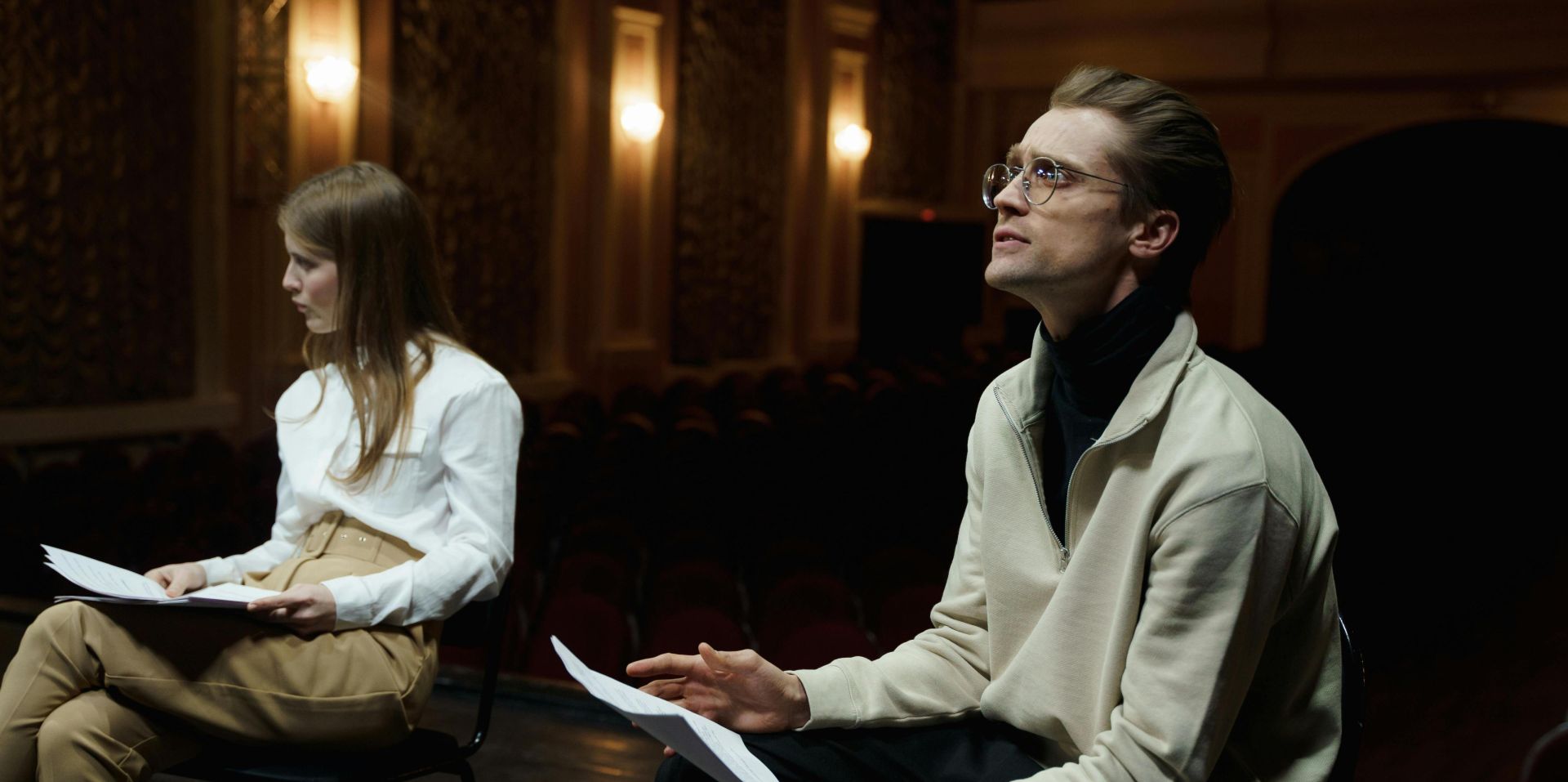 Two actors rehearsing with scripts in a dimly lit theater auditorium.