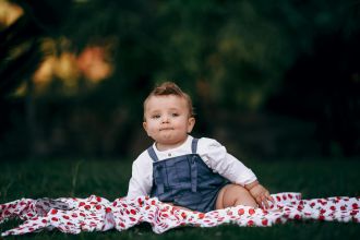 Charming baby boy sitting on a strawberry-patterned blanket in a lush garden setting. Captivating portrait.